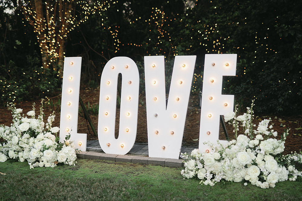 Oversized illuminated “LOVE” marquee letters surrounded by white wedding florals at an outdoor evening reception.