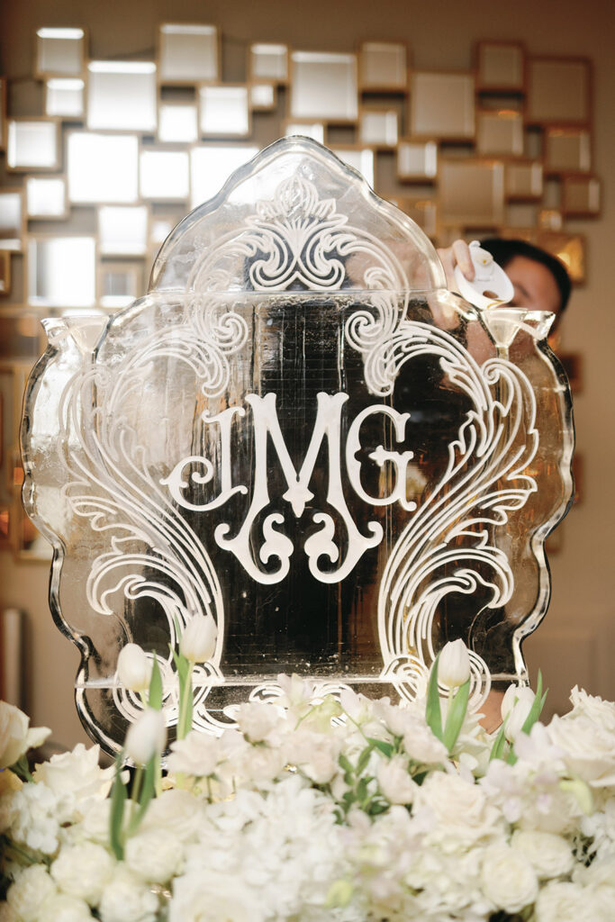 Hand-carved ice sculpture with engraved monogram surrounded by white florals at the wedding bar.