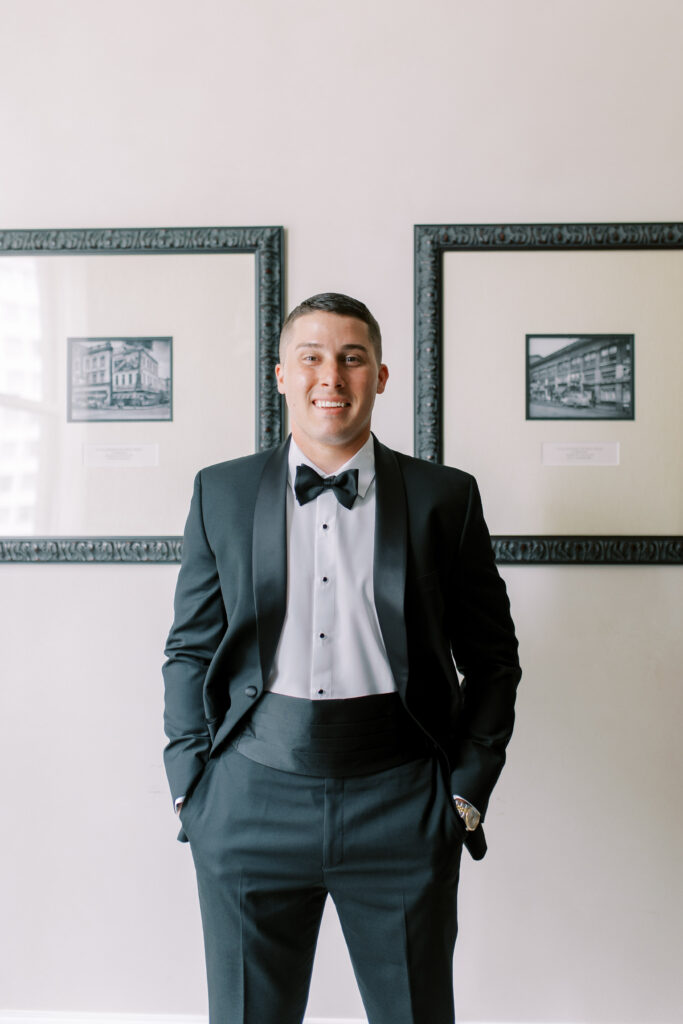 Groom posing in a classic black tuxedo with bow tie, photographed indoors against a neutral wall with framed artwork.