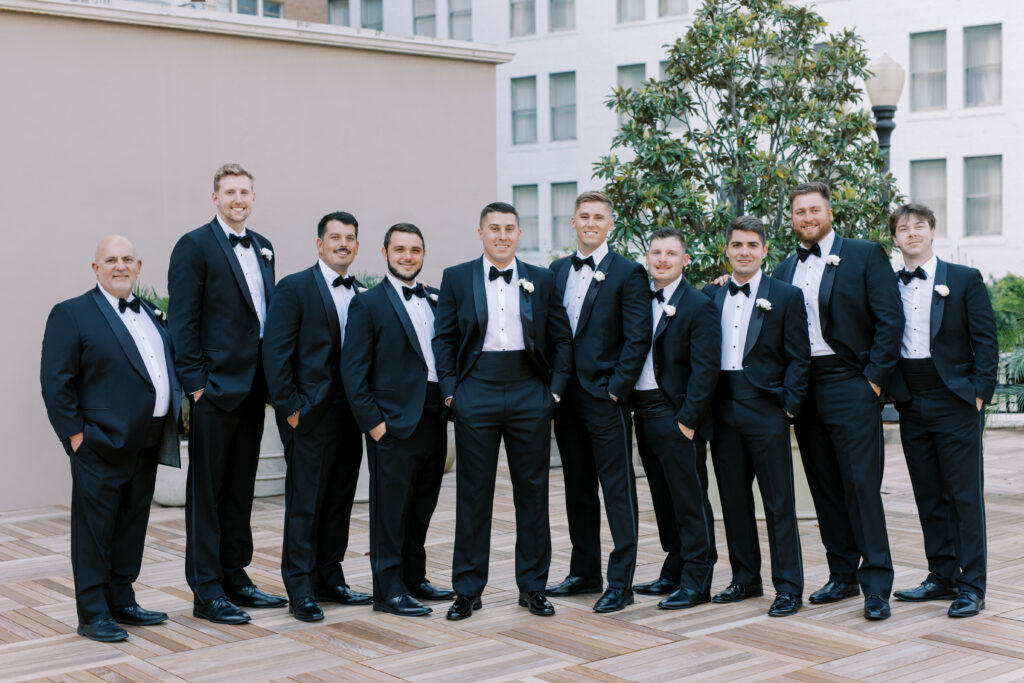 Groom and groomsmen lined up in black tuxedos on a rooftop terrace, captured in a formal wedding party portrait.