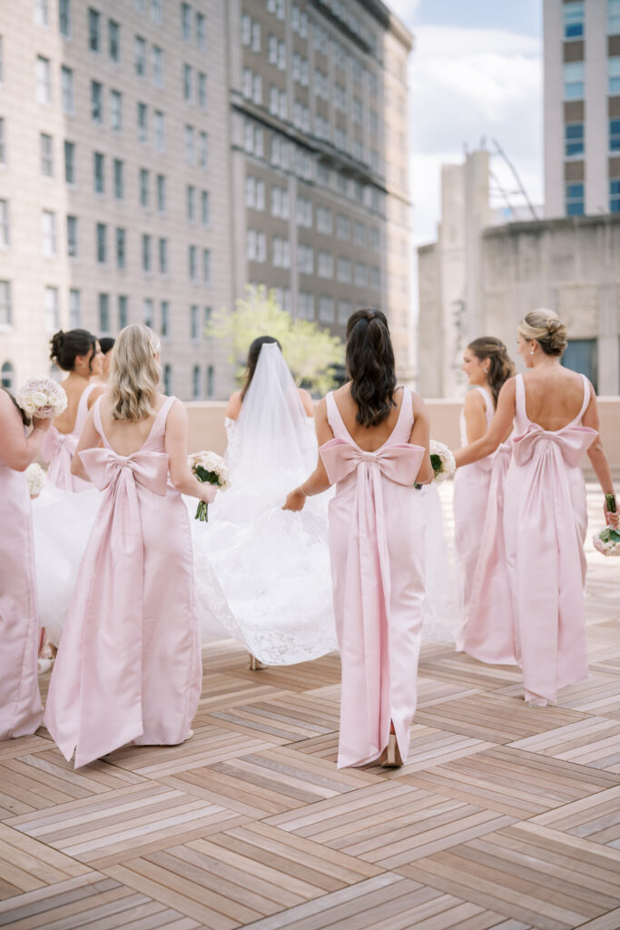 Bridesmaids in blush dresses carrying the bride’s train as they walk together on a rooftop terrace.