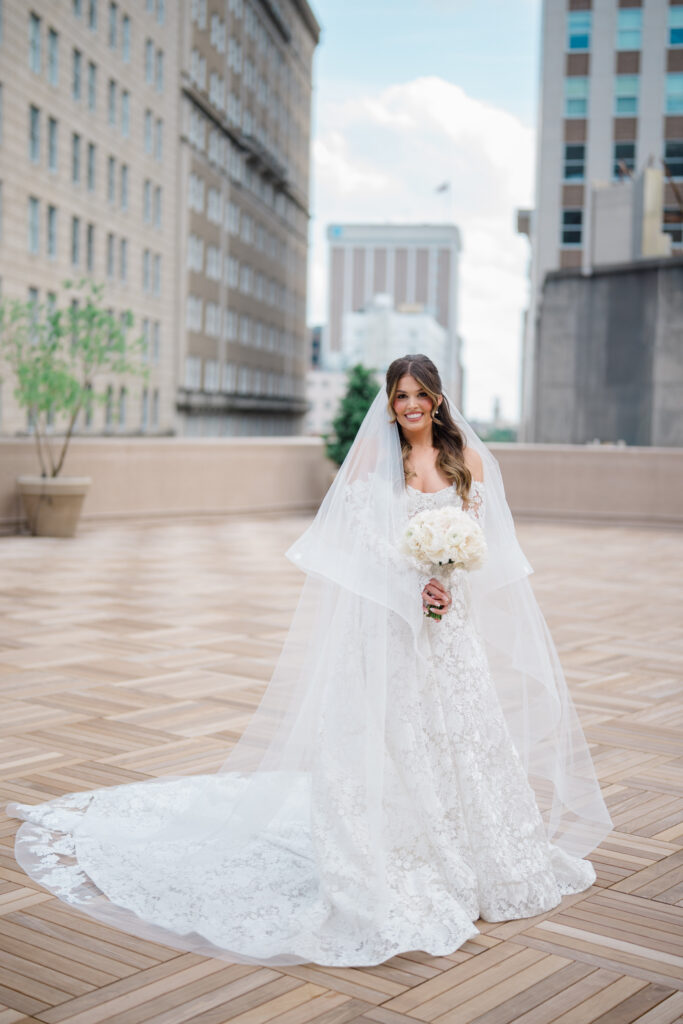 Bride holding a white rose bouquet and smiling beneath her veil during a rooftop portrait with the city skyline behind her.