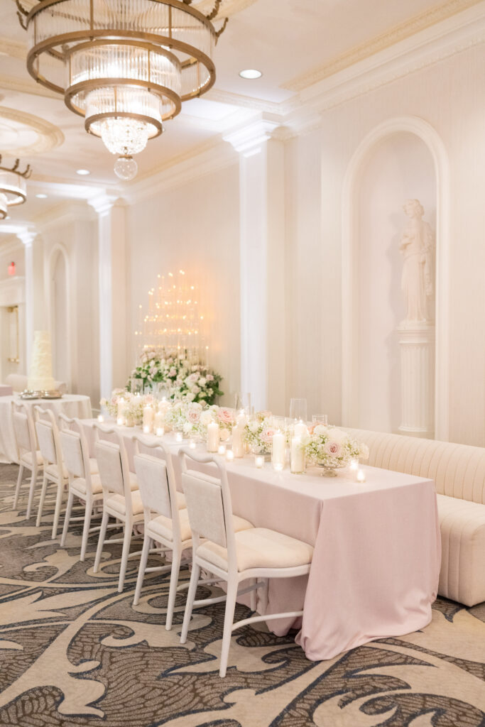Long banquet table styled with blush linens, garden florals, and candlelight beneath ornate chandeliers.