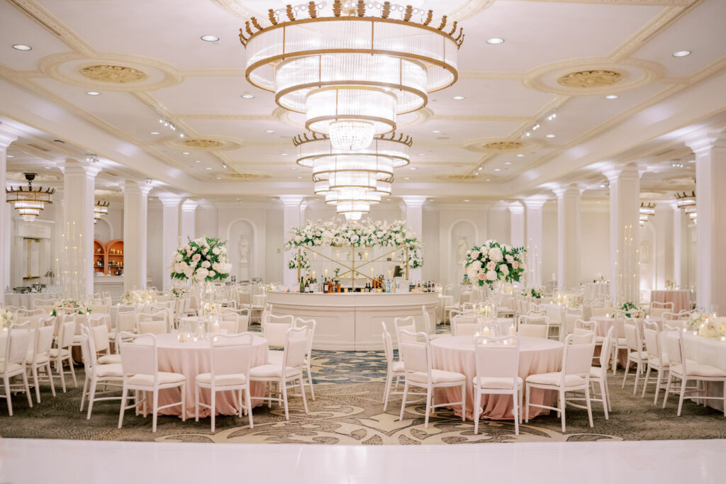 Grand ballroom reception reveal featuring round tables, blush linens, white chairs, and a central floral-adorned bar beneath chandeliers.