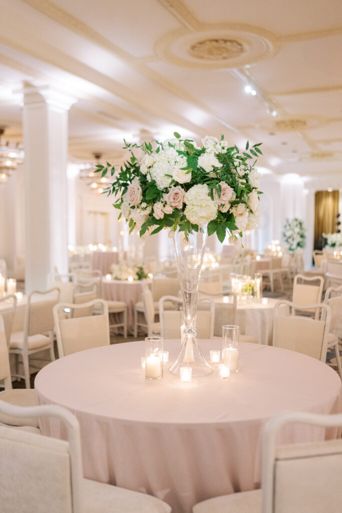Tall glass floral centerpiece with white hydrangeas, blush roses, and greenery surrounded by glowing votive candles.