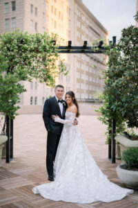 Bride and groom posing beneath a greenery-covered pergola on an outdoor terrace with the city skyline in the background.