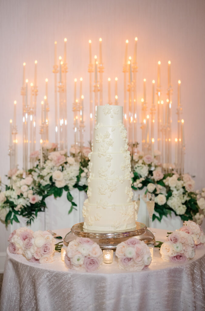 Five-tier white wedding cake with delicate floral detailing displayed on a round table surrounded by blush rose arrangements and candlelight.