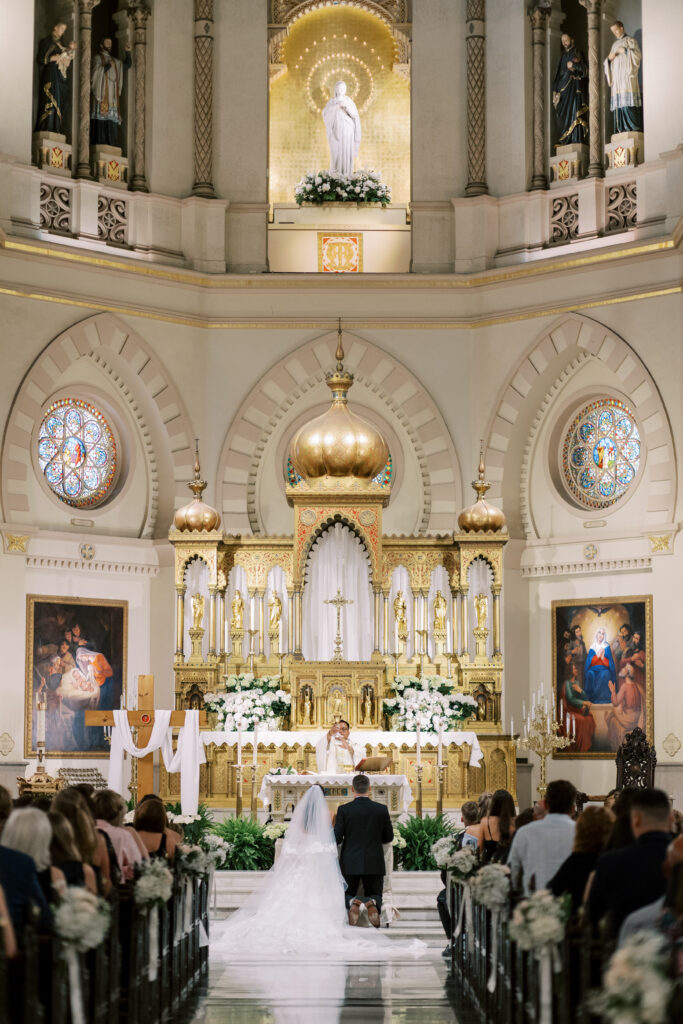 Bride and groom kneeling at the altar during a traditional church ceremony framed by gilded details and floral arrangements.