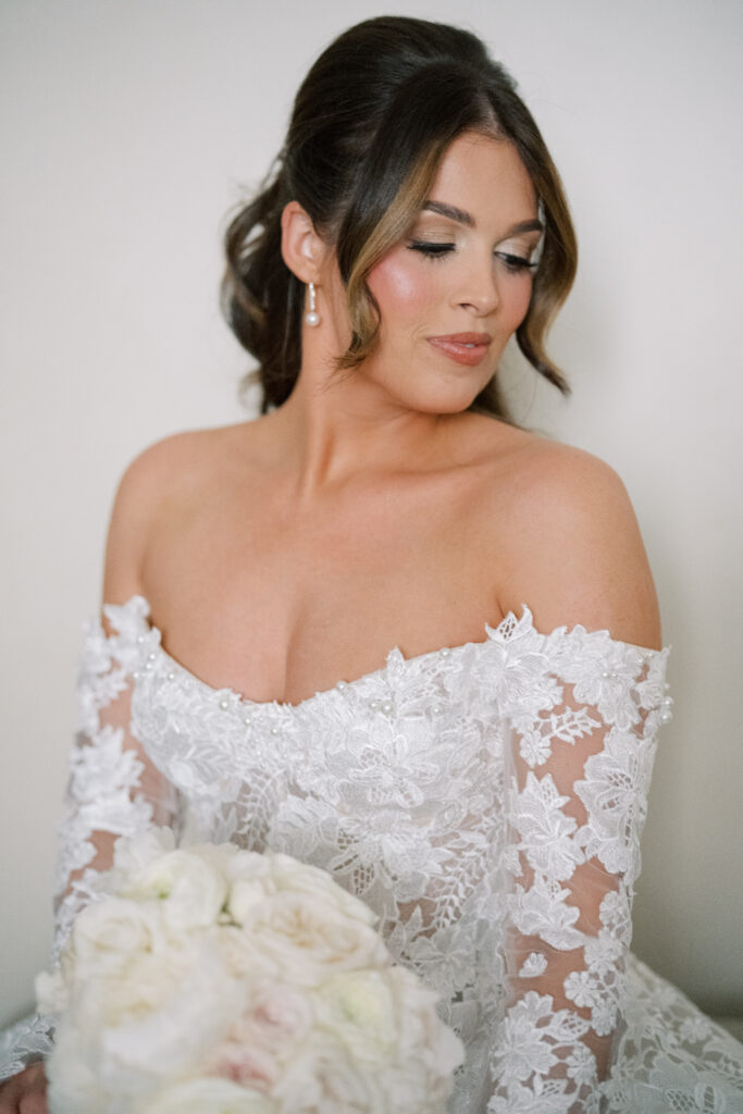 Soft bridal portrait highlighting the bride’s off-the-shoulder lace gown, pearl earrings, and natural makeup.