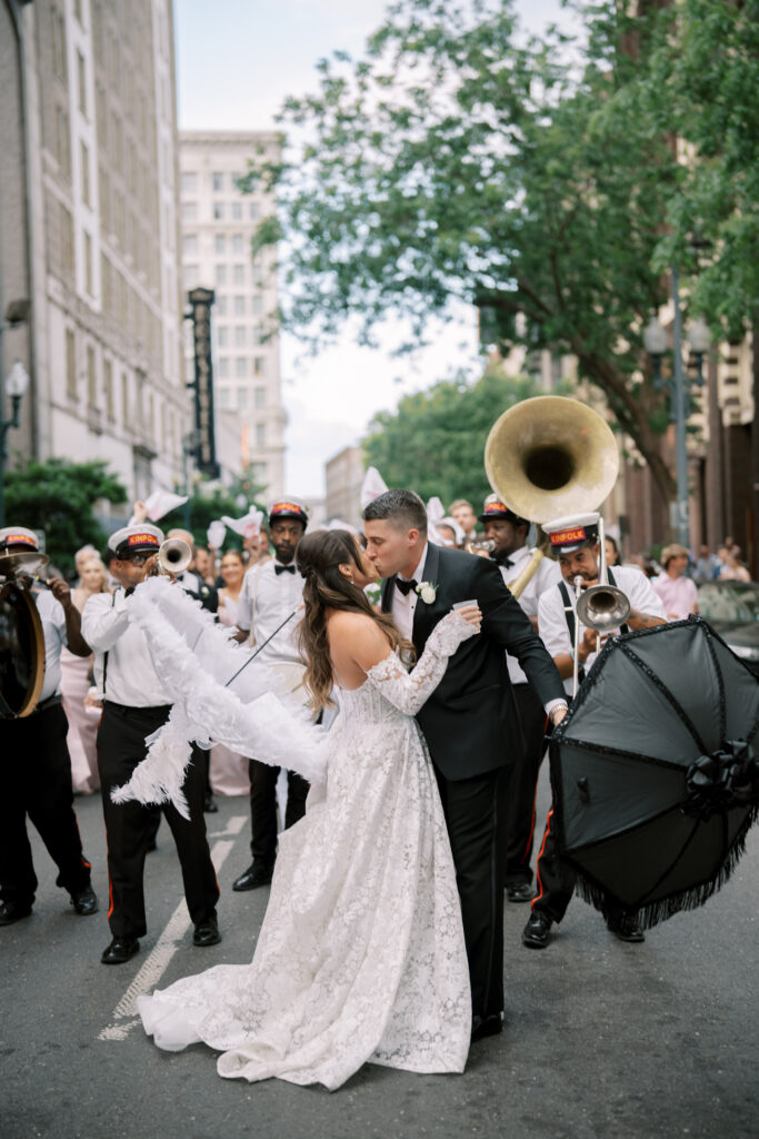 Bride and groom kissing during a lively New Orleans–style second line parade, surrounded by a brass band and guests.