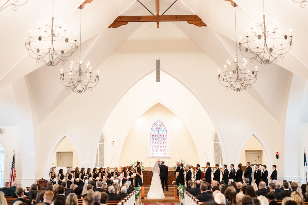 Wide ceremony view inside a bright chapel with arched ceilings, chandeliers, and the wedding party gathered at the altar.