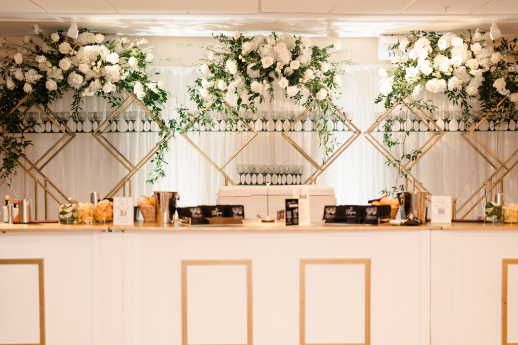 Elegant wedding bar with white paneled front, gold shelving, and abundant white florals cascading above glassware.