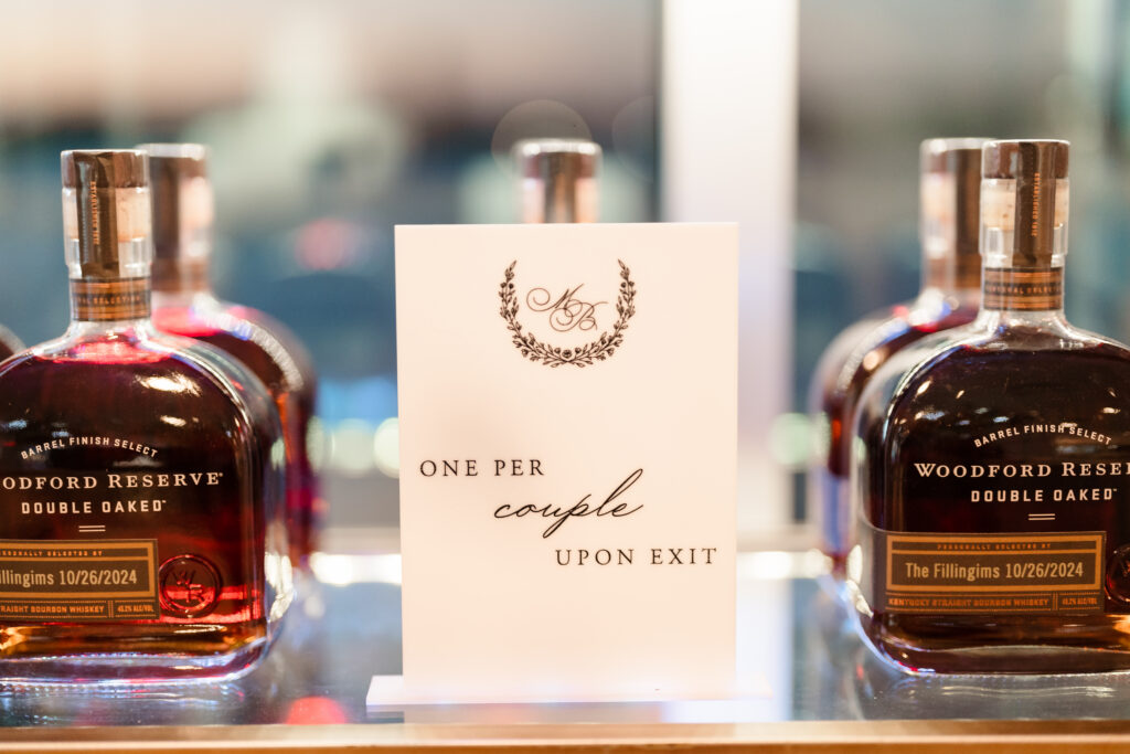 Personalized bourbon favors displayed with a calligraphy sign reading “One per couple upon exit,” flanked by engraved bottles.