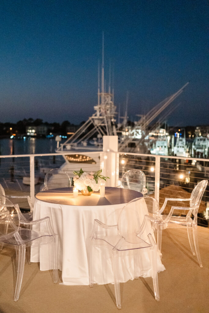 Intimate candlelit reception table set on a waterfront terrace with clear chairs and floral centerpiece at dusk.