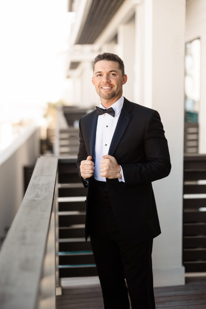Groom in a classic black tuxedo posing confidently along a sunlit outdoor walkway before the ceremony.