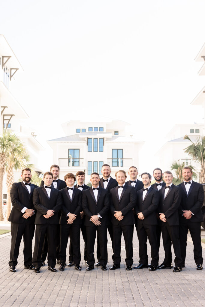 Groom and groomsmen in matching black tuxedos standing together in a formal outdoor group portrait.