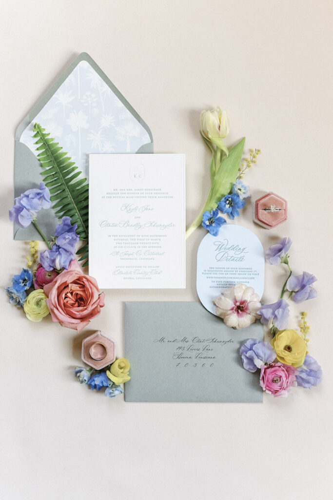Pastel wedding invitation suite styled flat-lay with spring florals, ring box, and soft blue accents.
