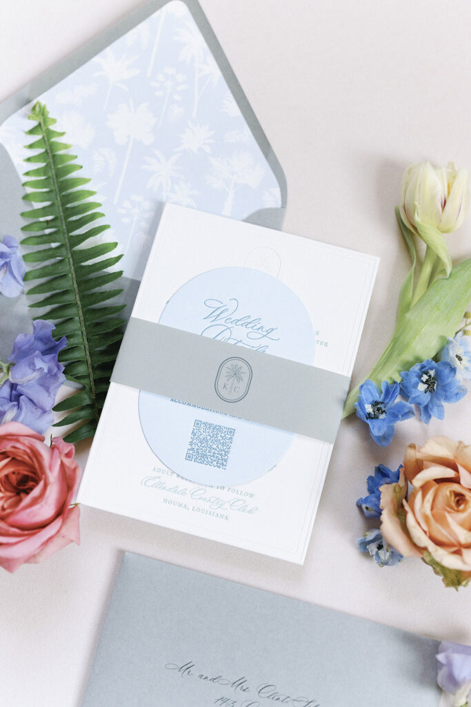 Close-up of invitation suite featuring a custom monogram band and QR code, styled with fresh blooms.