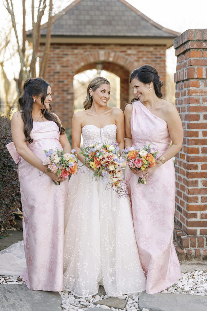 Bride posing with bridesmaids in blush dresses, all holding colorful bouquets in front of brick columns.