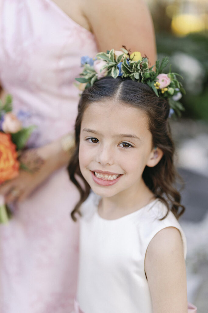 Smiling flower girl wearing a floral crown and white dress, captured in a soft outdoor portrait.