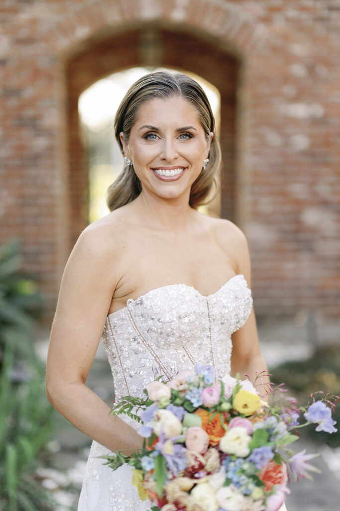 Bride holding a colorful spring bouquet, standing beneath a brick archway in natural light.