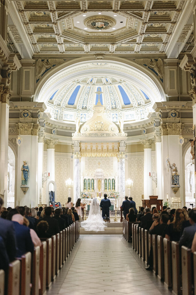 Wide ceremony view inside a grand church with an ornate domed ceiling and guests seated in pews.