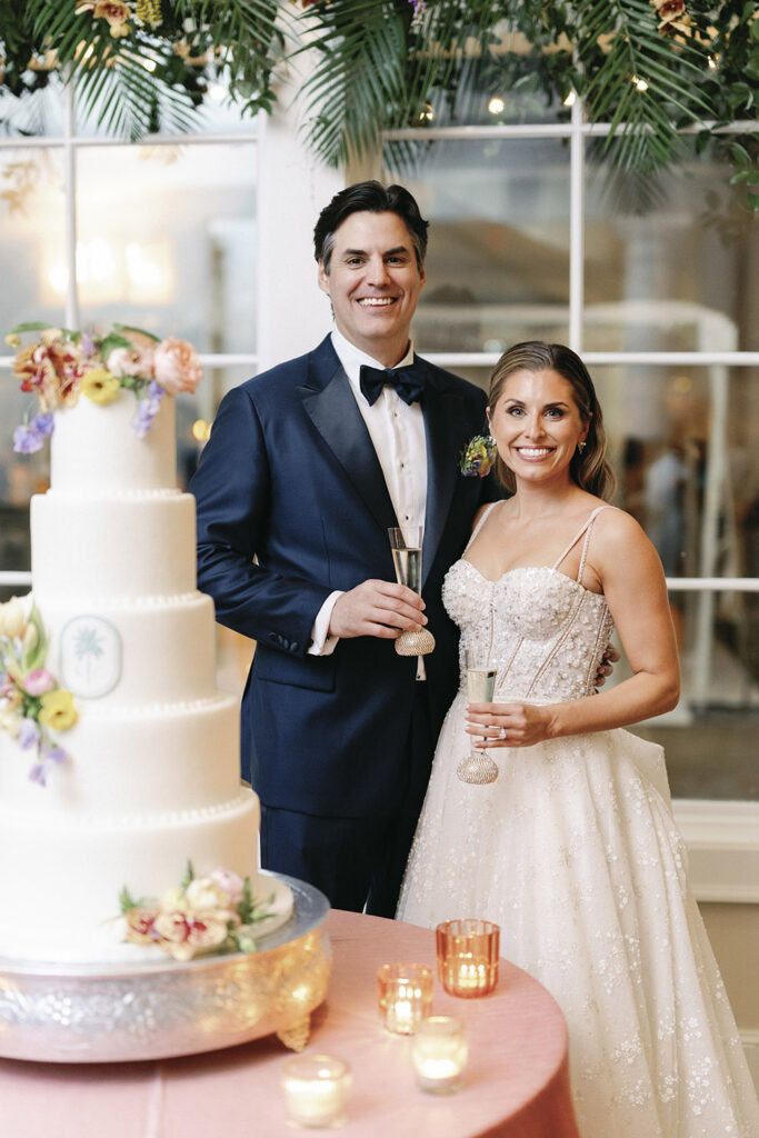 Bride and groom toasting beside their wedding cake, holding champagne flutes and smiling during the reception.