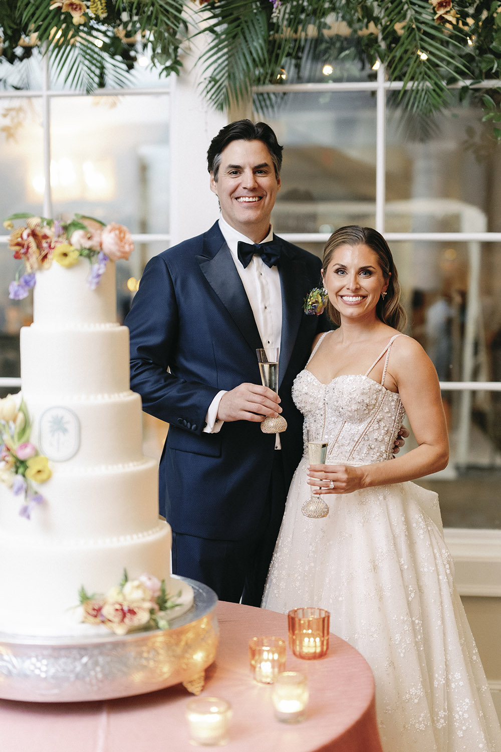 Bride and groom toasting beside their wedding cake, holding champagne flutes and smiling during the reception.