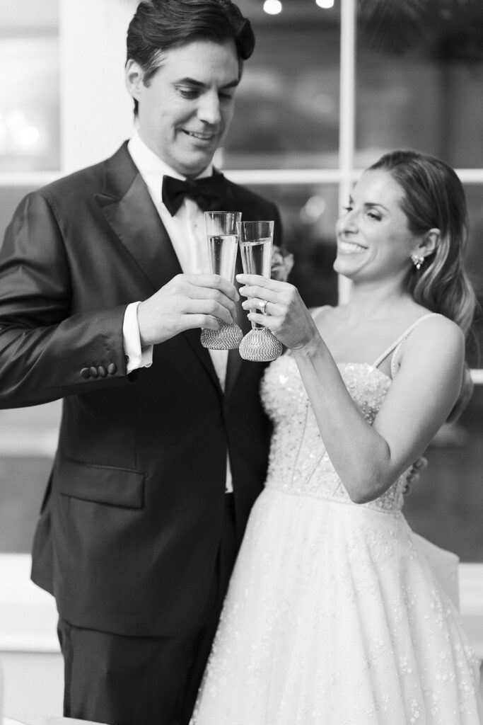 Black-and-white portrait of the bride and groom clinking champagne glasses in a celebratory toast.