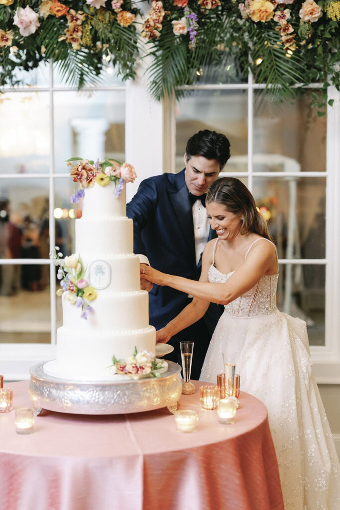 Bride and groom cutting their wedding cake together beneath floral greenery and warm candlelight.