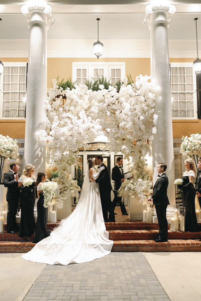 Bride and groom share a kiss beneath a dramatic white floral ceremony arch framed by grand columns at night.