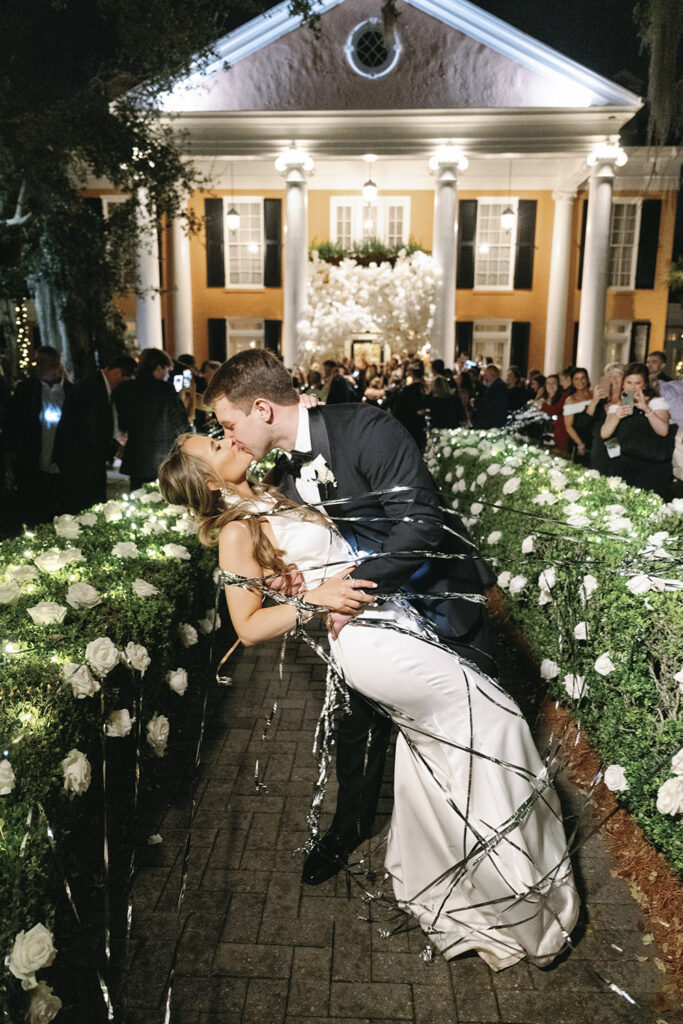 Bride and groom sharing a romantic dip kiss during their nighttime wedding exit with string lights and floral-lined walkway.
