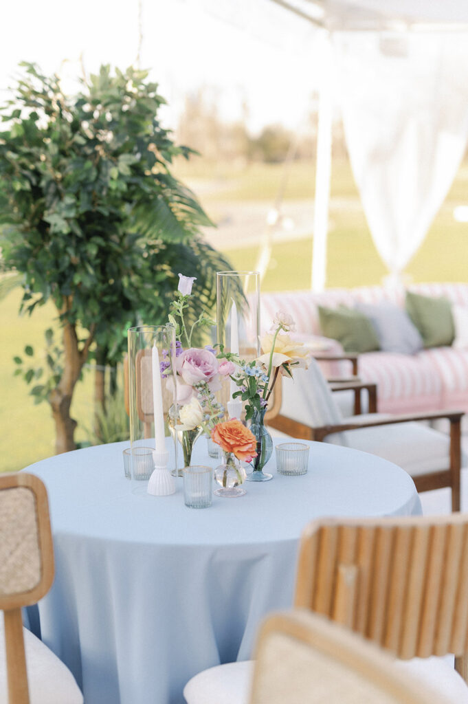 Pastel lounge vignette with a blue cocktail table styled with soft floral arrangements and glass candle holders under a clear-top tent.