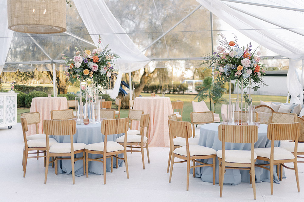 Elegant reception tables featuring pale blue linens, light wood chairs, and lush floral centerpieces beneath a draped clear tent.