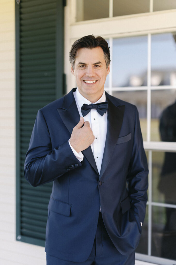 Groom in a classic navy tuxedo adjusting his bow tie, standing outside a white-paneled venue with green shutters.