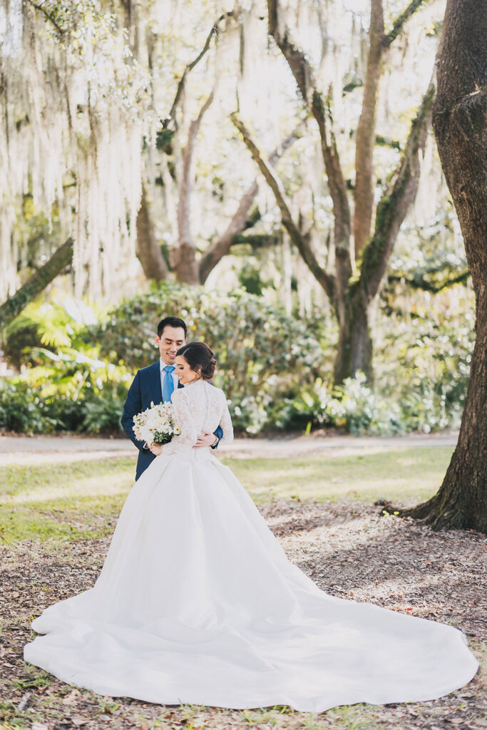 Bride and groom embracing beneath towering oak trees, showcasing the flowing train of the bride’s gown.