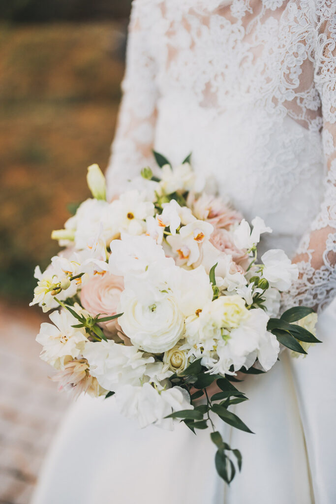 Close-up of the bride’s white floral bouquet with ranunculus and greenery against her lace wedding gown.