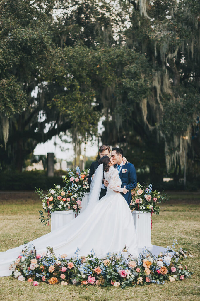 Bride and groom sharing their first kiss at the altar during an outdoor ceremony framed by lush florals and oak trees.