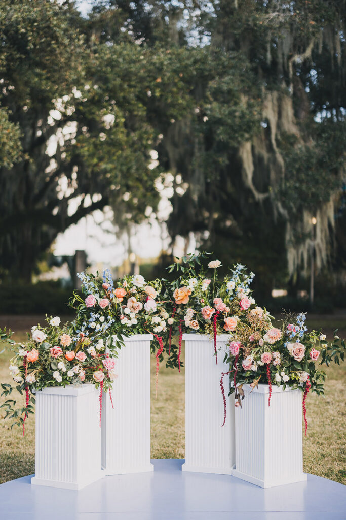 Outdoor ceremony altar styled with white pedestals overflowing with pastel garden florals beneath moss-draped oak trees.