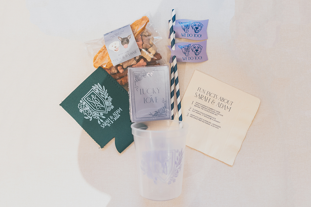 Wedding welcome bag flat lay featuring custom koozie, dog treats, cocktail stirrers, and printed cards with playful illustrations.