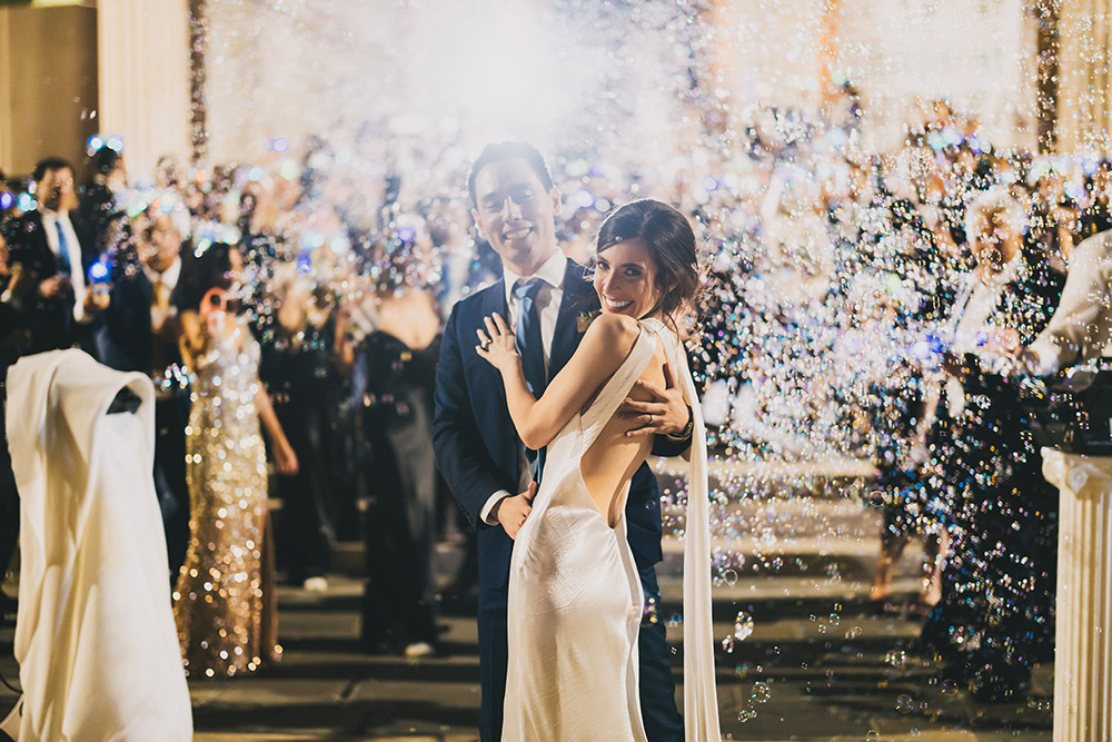 Bride and groom embracing during their bubble-filled exit at night, surrounded by cheering guests outside the venue.