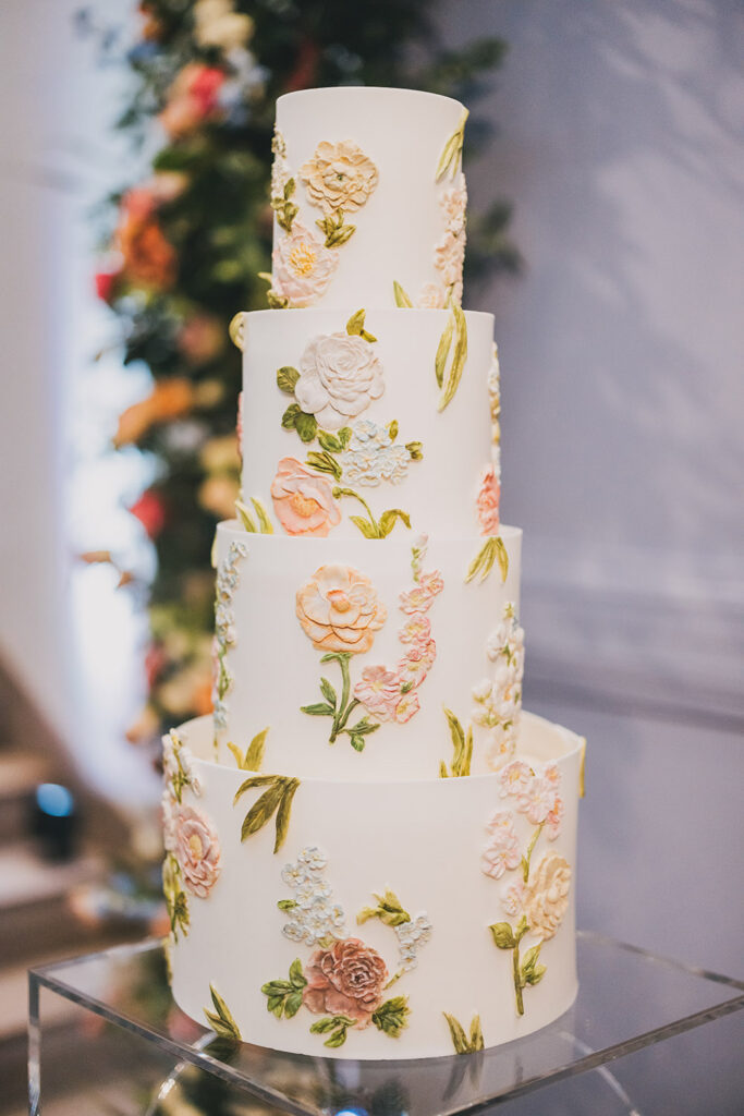 Four-tier white wedding cake decorated with hand-painted pastel florals displayed on a clear pedestal.