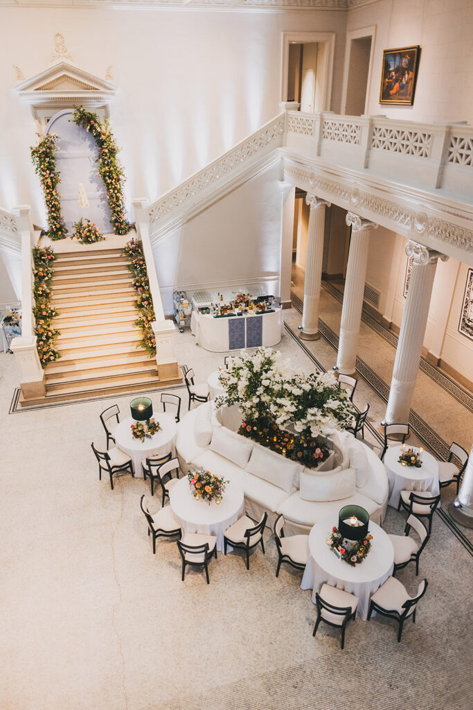 Overhead view of the museum reception lounge showcasing round tables, white seating, and a floral-filled staircase backdrop.