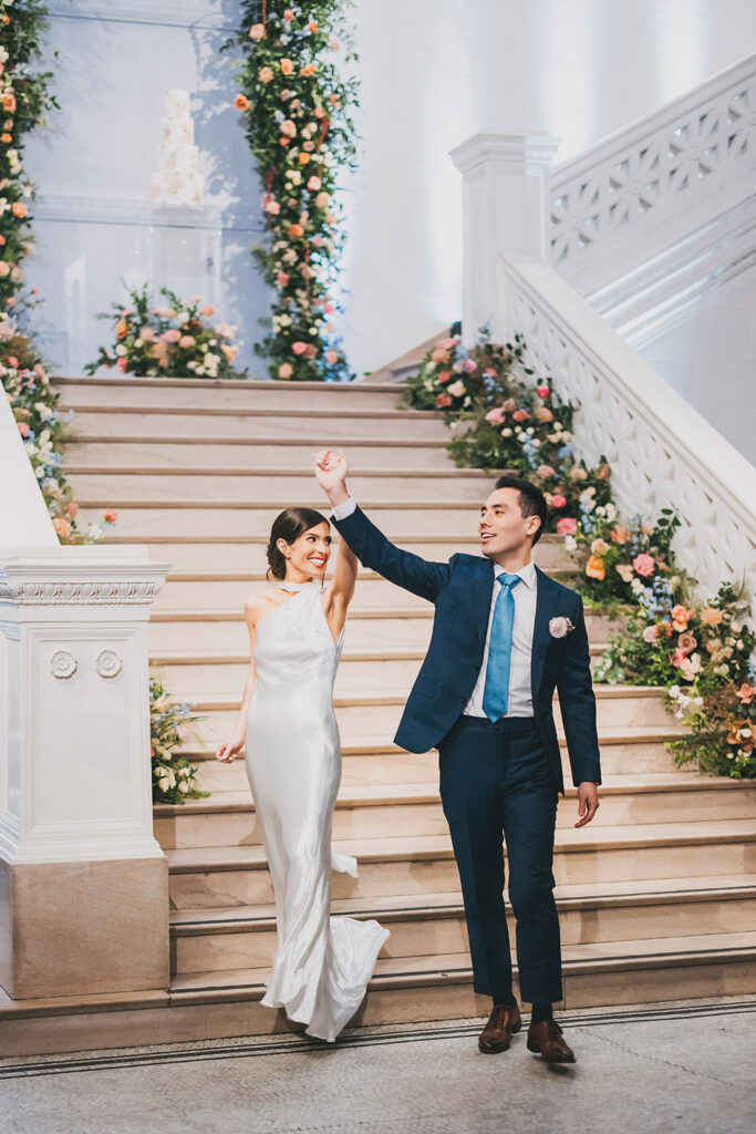 Bride and groom making a joyful grand entrance down a grand staircase adorned with cascading florals inside a museum reception space.