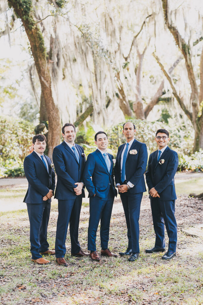 Groom and groomsmen standing together outdoors in navy suits beneath oak trees, captured in a classic wedding party portrait.