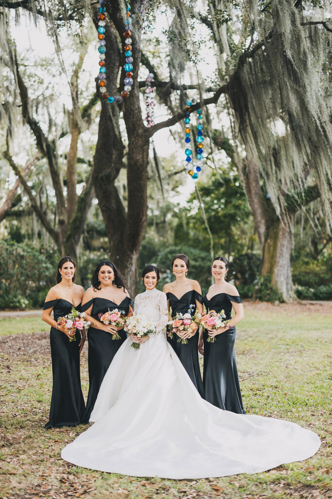 Bride posing with her bridesmaids in sleek black dresses beneath moss-draped oak trees, holding colorful garden-style bouquets.