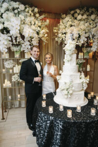 Bride and groom standing beside a towering white wedding cake adorned with florals and candlelit décor.