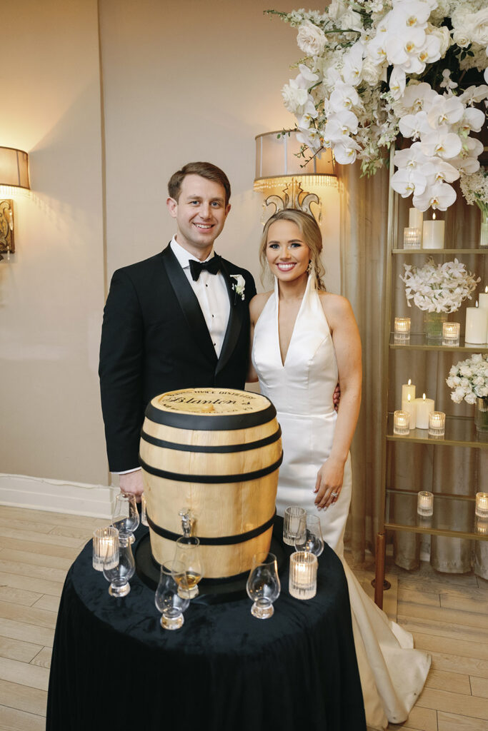 Bride and groom smiling beside a personalized whiskey barrel display at an elegant wedding reception.