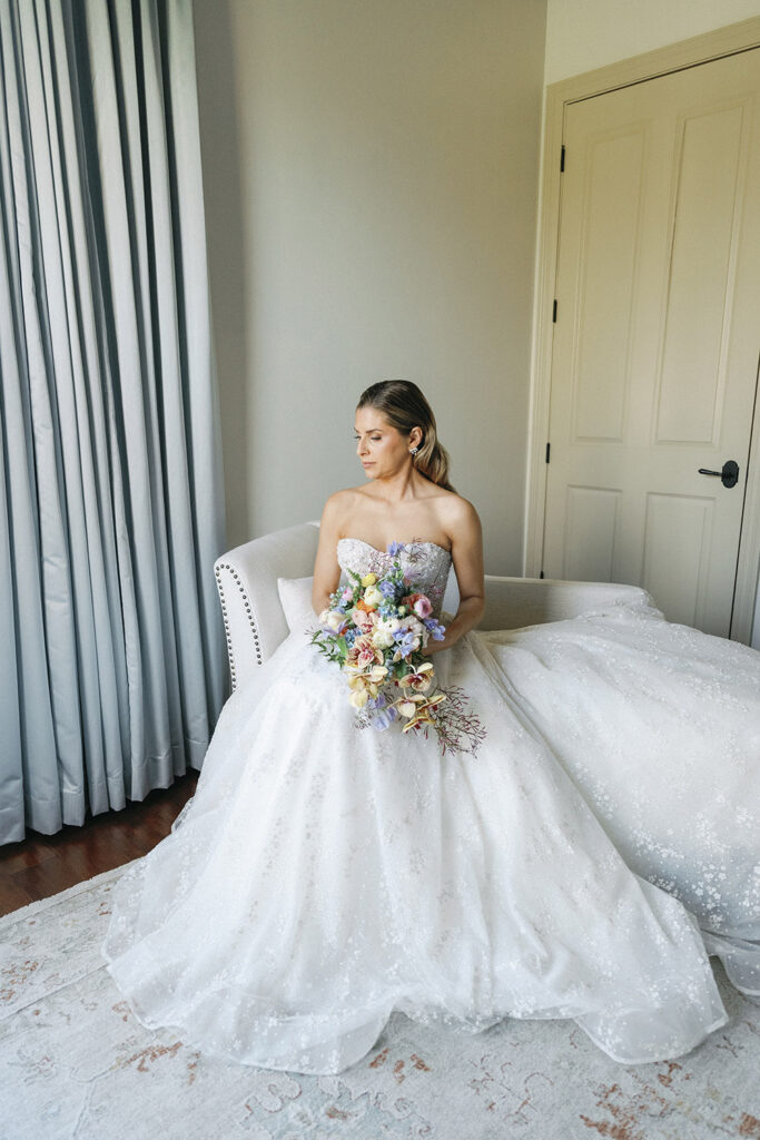 Bride seated on a cream sofa holding a colorful garden-style bouquet, wearing a strapless ball gown in a softly lit room.