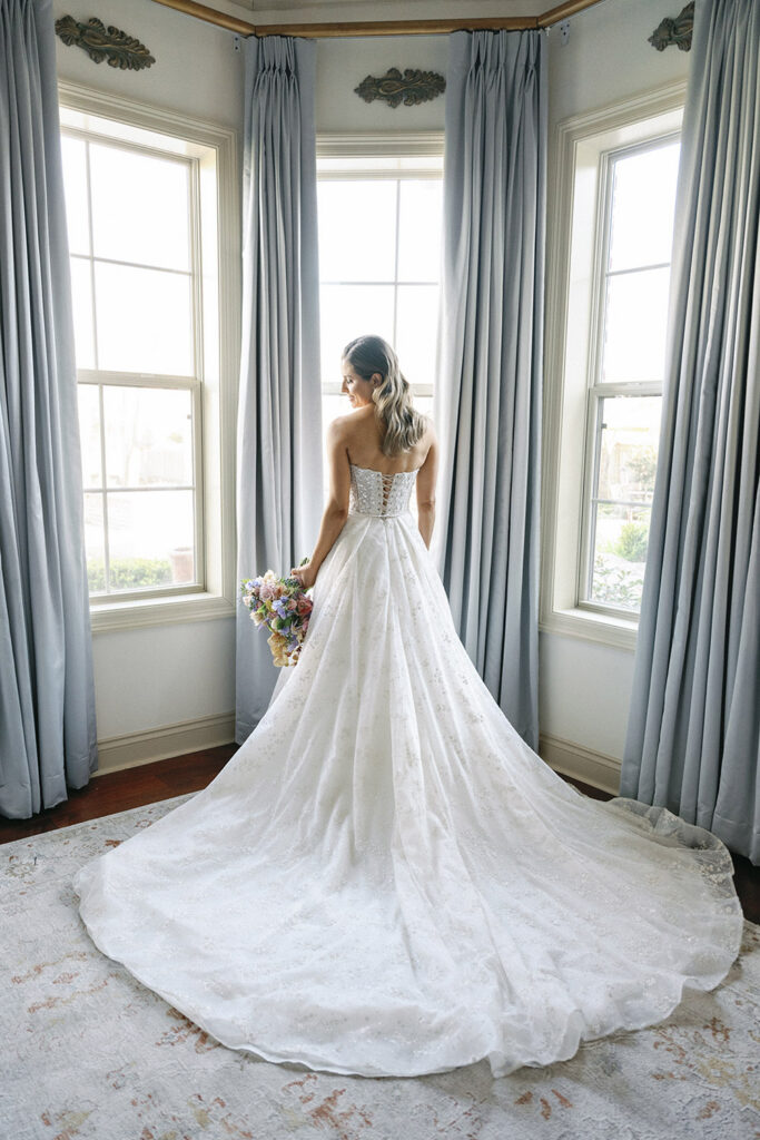 Back view of the bride standing between tall windows, showcasing the flowing train of her wedding gown and pastel bouquet.